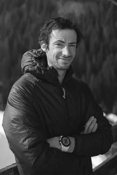 Kilian Jornet posing in the mountains with his APEX smartwatch for mountain and trail runners. Kilian Jornet wears a Grey down jacket and the background is dark with mountain trees. a